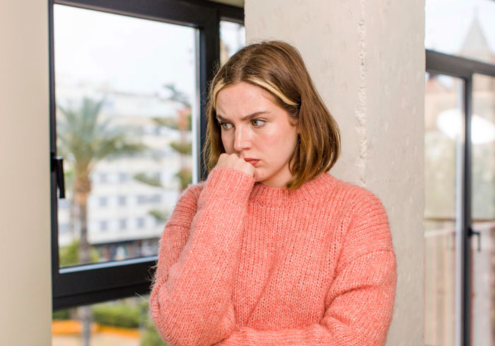 A woman in a pink sweater looks upset, standing by a window, reflecting on exclusion from a birthday party.