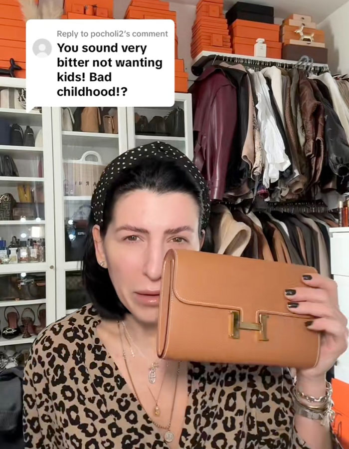 Millennial woman holding a designer clutch in a closet, responding to a critical comment about her child-free lifestyle choices.