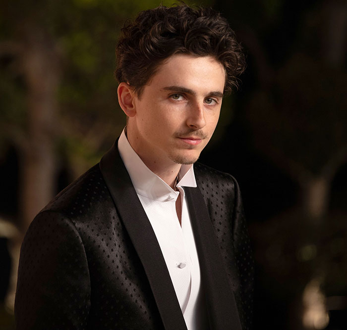 Timoth&eacute;e Chalamet wearing a black suit and white shirt, looking at the camera in an outdoor setting.