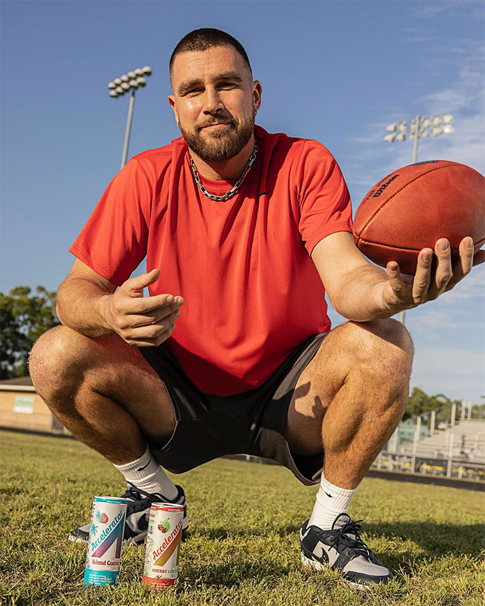 Travis Kelce in a red shirt holding a football outdoors, with energy drinks on the grass, showcasing luxury lifestyle.