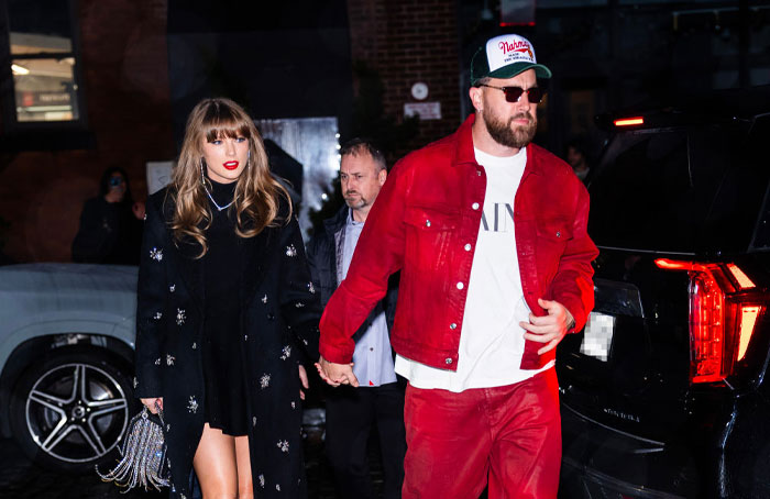 Travis Kelce in red jacket and hat with Taylor Swift in a black coat, walking by a luxury SUV at night, highlighting luxury lifestyle.