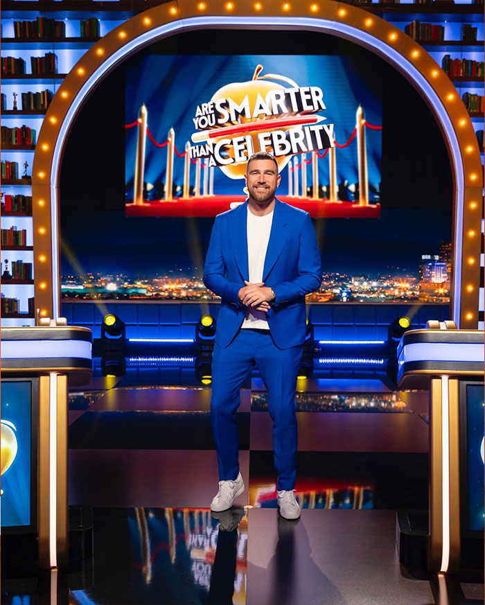 Travis Kelce in a blue suit on stage with a city backdrop, highlighting luxury lifestyle elements.