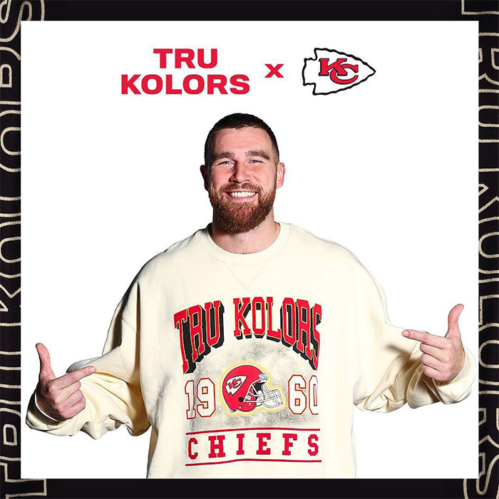 Travis Kelce in Tru Kolors Chiefs sweatshirt, smiling and pointing, representing luxury lifestyle off the field.