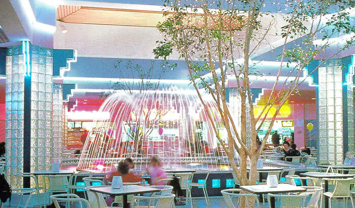 Vintage mall food court with neon lights, trees, fountain, and retro seating.