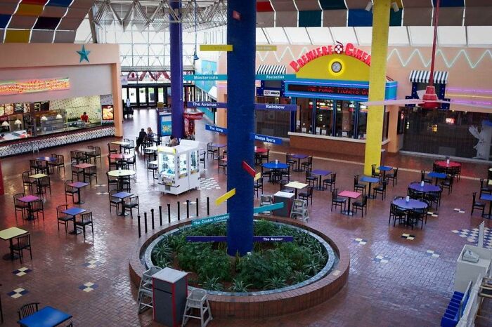 Vintage mall food court with colorful tables, retro design, and restaurants in the background.