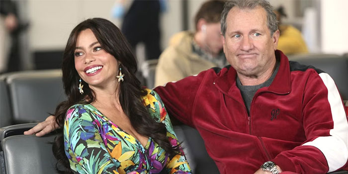 TV couple sitting on airport chairs, the man in a red jacket and the woman in a colorful dress, both smiling.