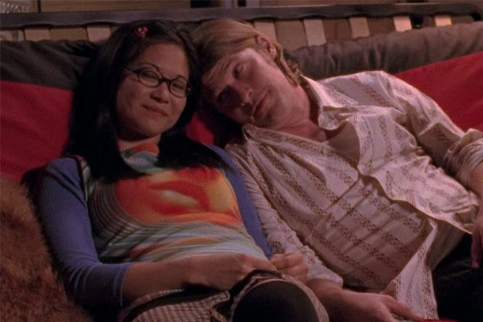 TV couple with less chemistry, sitting close on a couch, one wearing glasses and a patterned shirt, the other in a striped shirt.