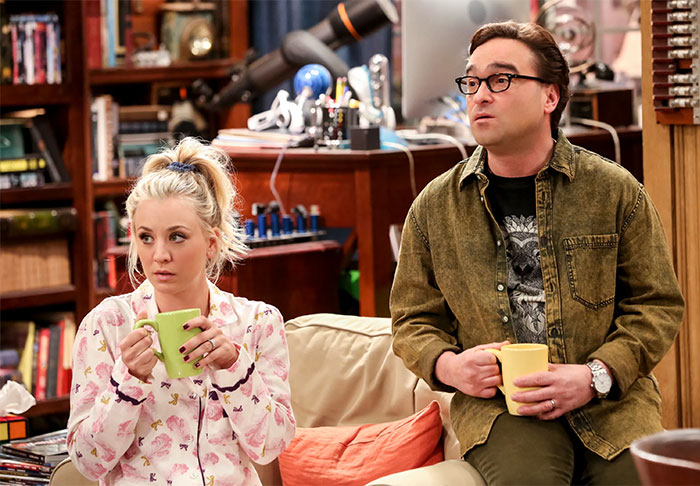 Two TV characters sitting on a couch, holding mugs, portraying a couple with less chemistry.