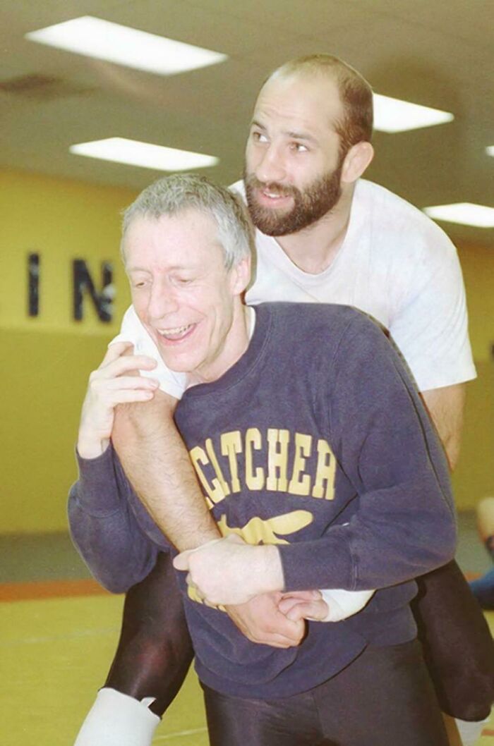 Two men smiling during a playful wrestling session indoors, one wearing a sweatshirt, capturing a moment with a creepy history.