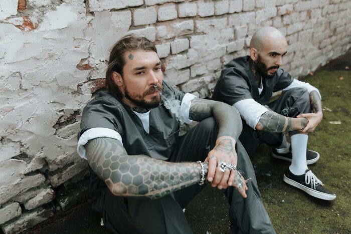Two men with tattoos sit against a brick wall, possibly undercover cops in a tense situation.