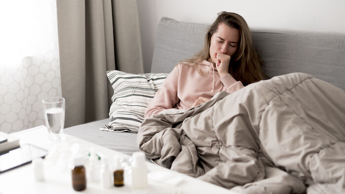 A woman in bed looking unwell, surrounded by unexpected things like medicine bottles and tissues.