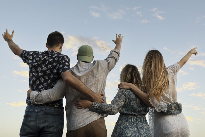 Four friends with arms around each other, facing a clear sky, symbolize reality's unexpected joys.