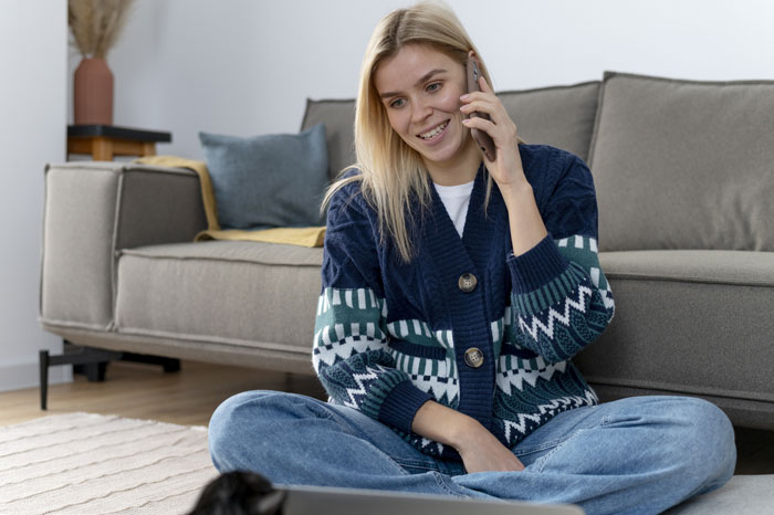 Woman on phone, thinking about friends' unruly kids, sitting on floor near sofa, wearing a blue patterned cardigan.