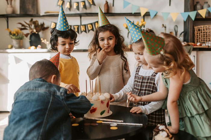 Children wearing party hats gather around a cake at a birthday celebration indoors.