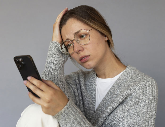 Woman wearing glasses looks upset while reading her phone, flabbergasted by canceled date plans.