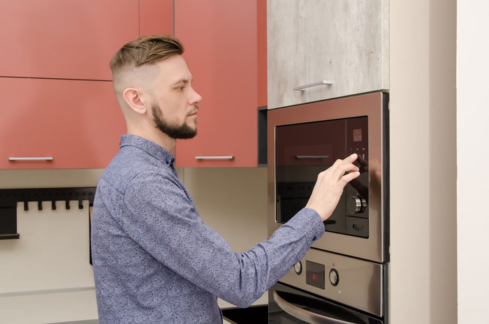 A man in a kitchen operating a microwave, wearing a patterned blue shirt, focused on heating breast milk.