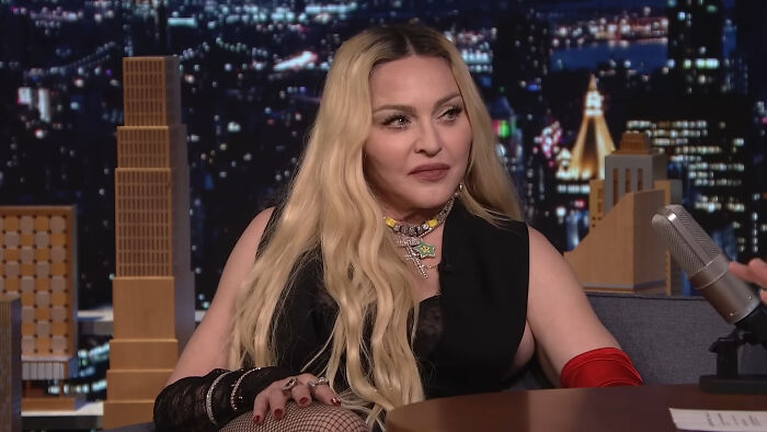 Celebrity with long blonde hair and red gloves on a talk show set, highlighting petty moments.