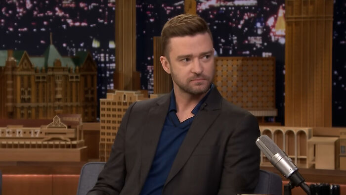 Celebrity sitting in a studio, wearing a dark suit and blue shirt, during a late-night talk show.