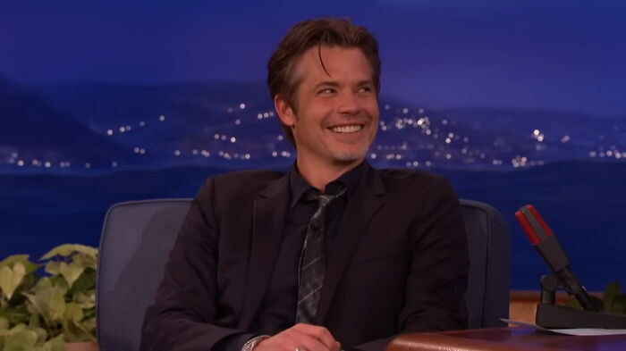 A smiling celebrity sits on a talk show set, wearing a dark suit, with city lights in the background.