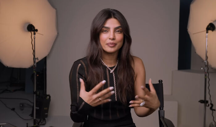 Priyanka Chopra talking energetically in a studio, seated with lighting equipment in the background.