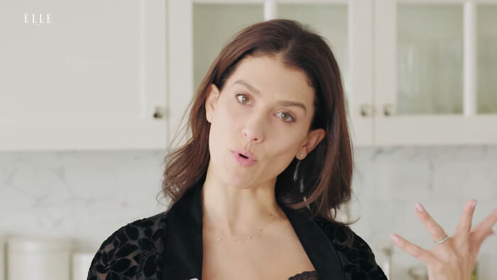 Woman in a kitchen with long hair, wearing a black top, gesturing while speaking. 