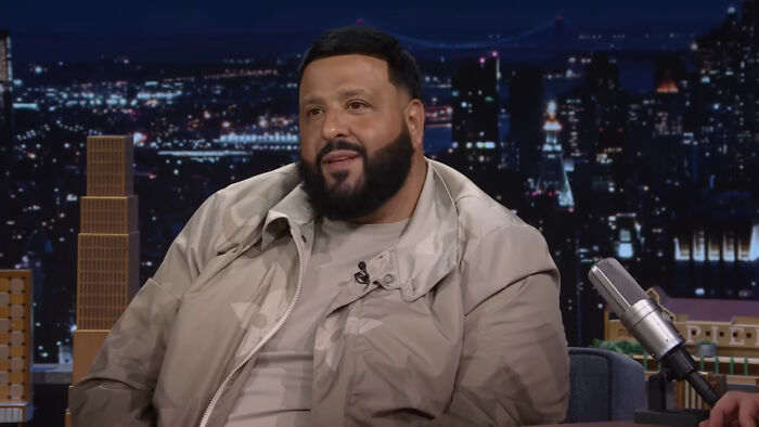 A person speaking on a late-night show set, wearing a beige jacket, with city skyline in the background. SEO keyword: "McGregor".