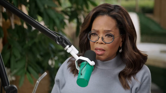 Oprah Winfrey person with glasses and a gray sweater speaking into a microphone, discussing celebrity conspiracy theories.