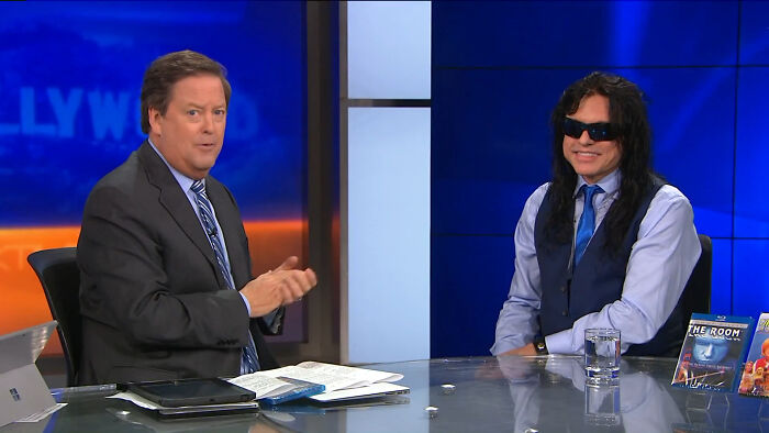 Two men discussing on a talk show, with DVDs on the table, related to celebrity conspiracy.