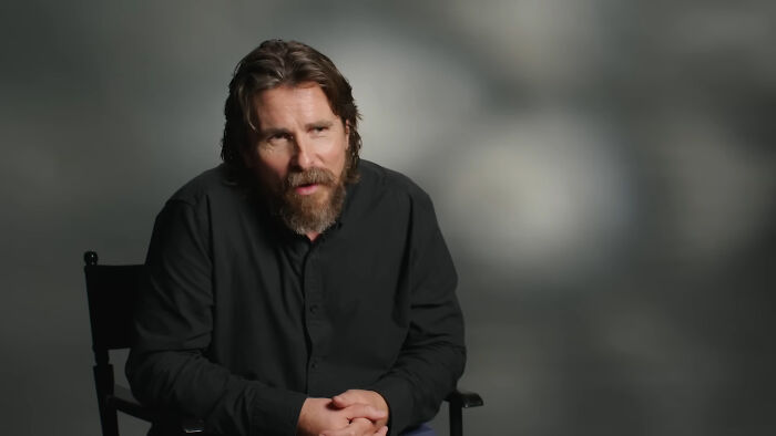 Man sitting and talking in an interview setting, wearing a black shirt. Celebrity conspiracy discussion backdrop.