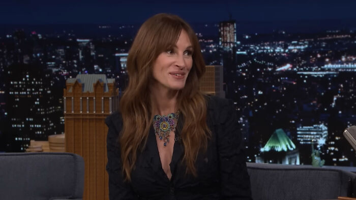 A woman with long hair in a black dress, smiling on a nighttime talk show set, cityscape backdrop.