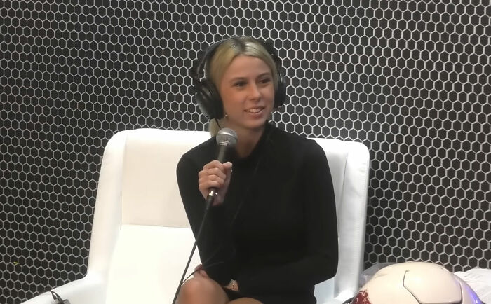 Person with headphones and mic, sitting on a white chair, casually dressed in black against a patterned backdrop at work.