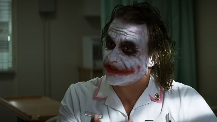 Actor in iconic role with white face paint and red smile, wearing a nurse outfit in a dimly lit room.