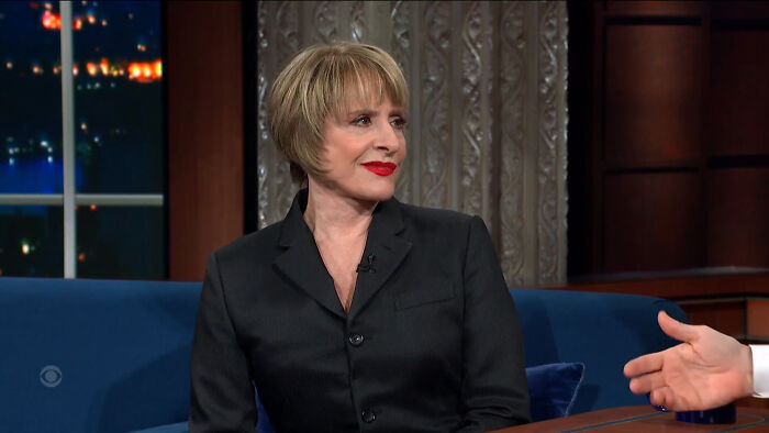 A celebrity in a black suit with short hair and red lipstick, sitting on a blue couch during a talk show.