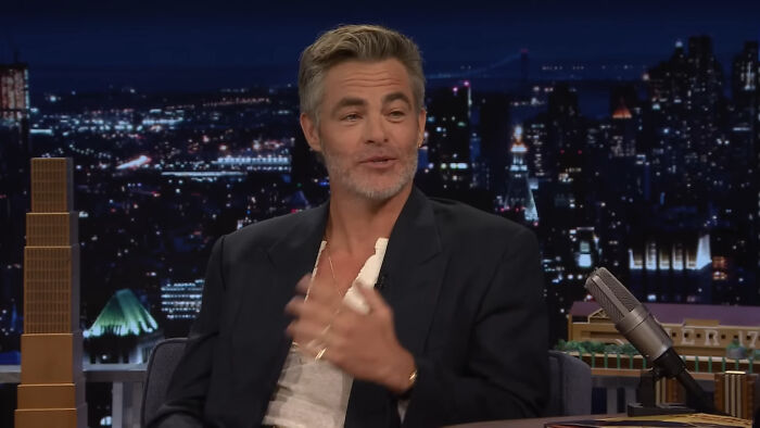 Brad Pitt in a suit gesturing on a talk show, city skyline backdrop, microphone visible; related to totally plausible celebrity conspiracy.