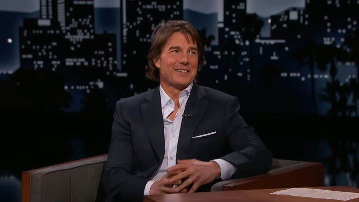 Man in a suit on a talk show set, smiling while discussing wild theories.