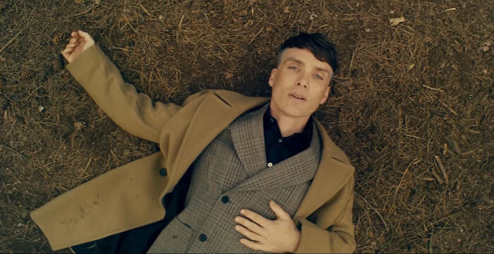 A person lying on the ground in a brown coat and gray suit, evoking celebrity charm.