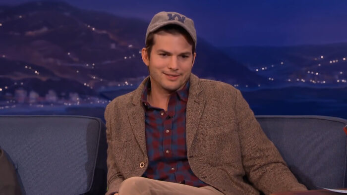 Ashton Kutcher in a cap and blazer on a talk show set, discussing a celebrity conspiracy topic.