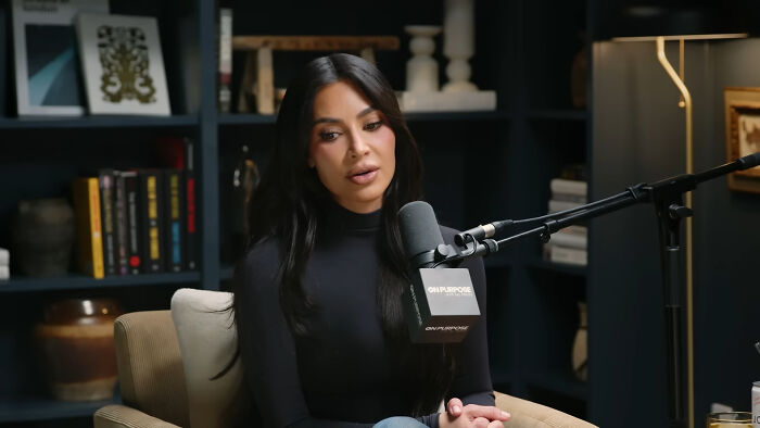 Kim Kardashian discussing plausible celebrity conspiracy on podcast, seated in a cozy room with bookshelves in the background.