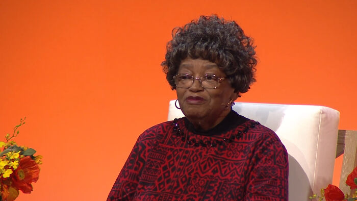 A woman in a red patterned dress sits on a white chair against an orange background, discussing historical facts.