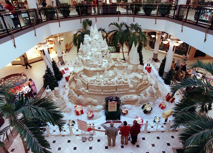 Vintage mall atrium with a large festive sand sculpture, surrounded by shoppers and palm trees.