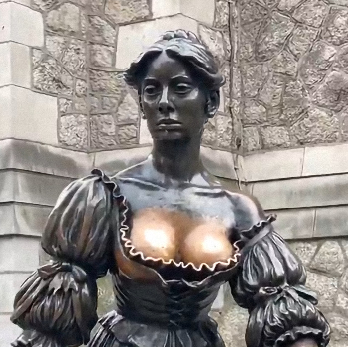 Statue of a woman with weathered bronze surface, wearing a historical dress, in front of a stone wall.