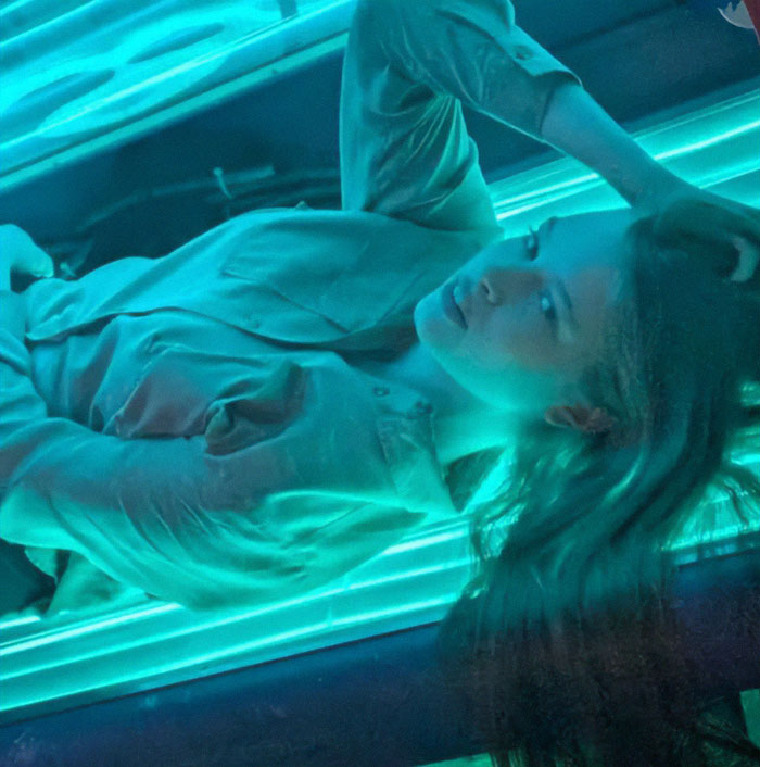 Woman under bright teal lights, lying down and looking upwards, reflecting an eerie atmosphere.