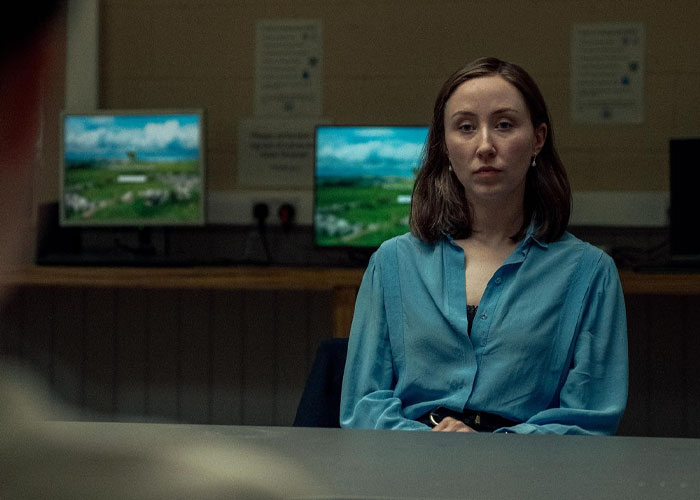 Woman in a blue shirt appearing serious, sitting in front of computers, related to TV show "Adolescence" and emoji meanings.