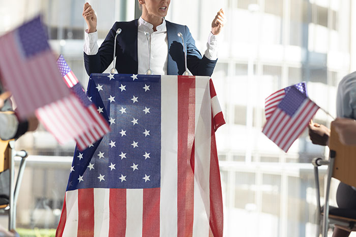 Speaker at podium with U.S. flag, addressing crowd waving American flags, highlighting what's normal in USA.
