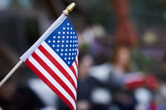 American flag on a small pole, held in front of a blurred outdoor background.