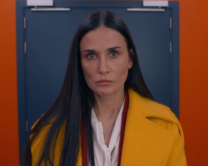 Demi Moore in a yellow coat, standing against a colorful backdrop.