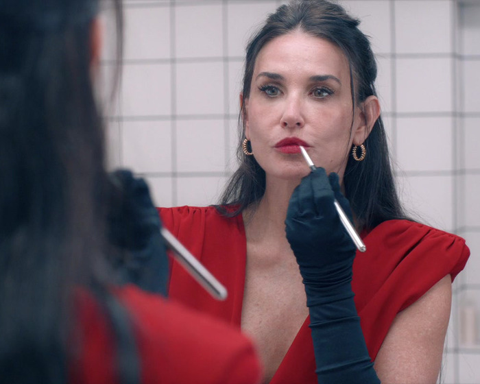 Demi Moore applying lipstick in a red outfit, looking in the mirror.