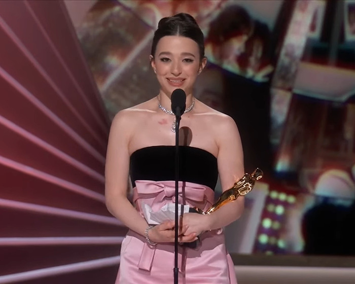 Mikey Madison holding an Oscar on stage, smiling in a pink dress.