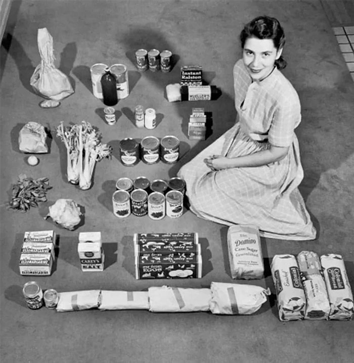 A woman in vintage clothing sits with 1950s American groceries arranged on the floor, showcasing everyday life in the past.