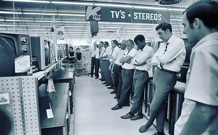 Men in a vintage electronics store, browsing TVs and stereos, depicting past everyday American life.